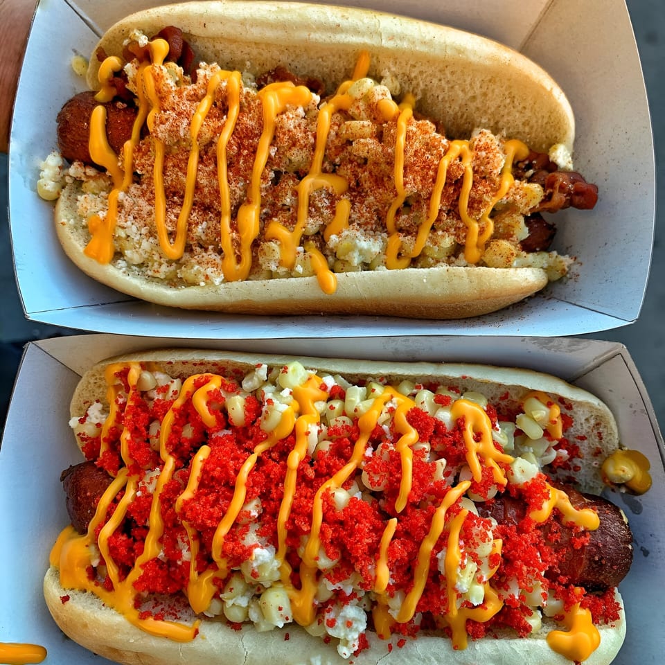 Two hot dogs in buns, one with mac and cheese, the other with corn and red crumbs.